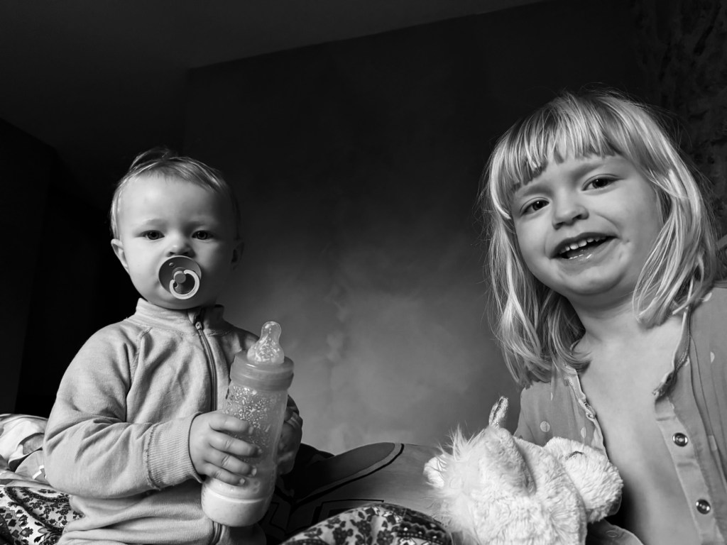 Ett litet barn med napp och nappflaska i handen sitter bredvid ett annat barn som ler, medan de är omgivna av en mysig inredning.