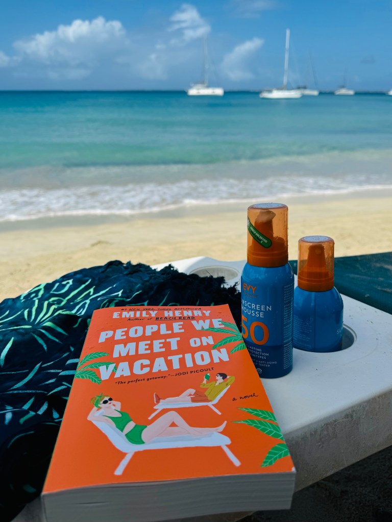 En bok med titeln 'People We Meet on Vacation' av Emily Henry ligger på en strandhandduk med havet i bakgrunden, tillsammans med två solskyddsprodukter.