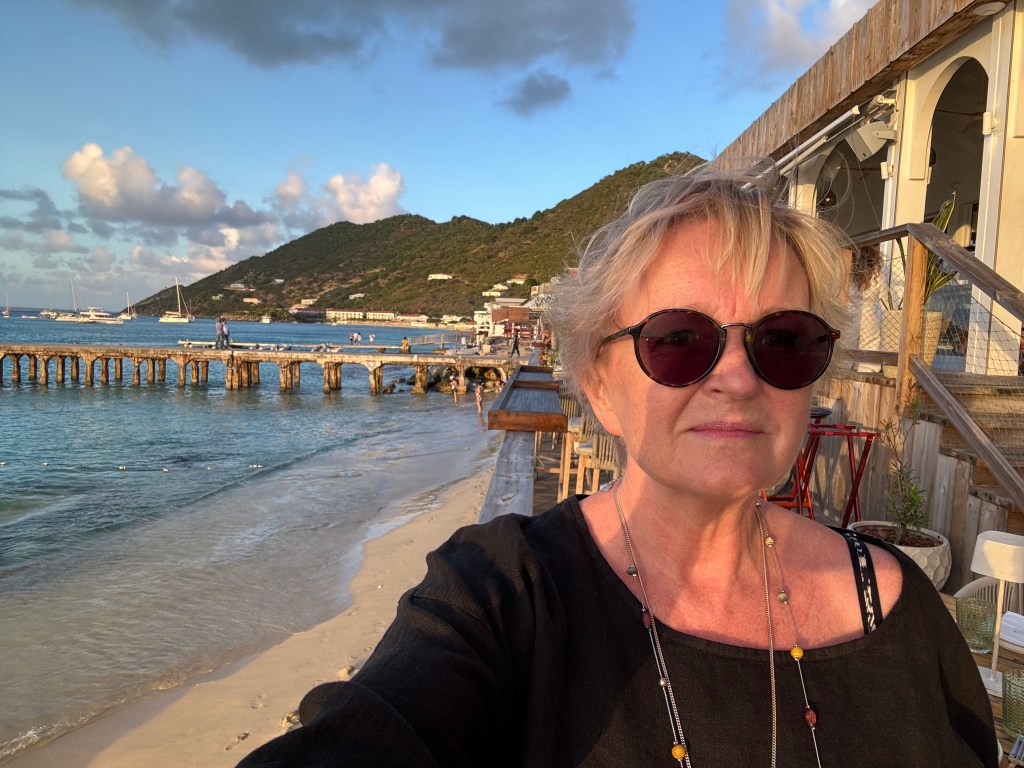 En kvinna med solglasögon står vid stranden, med en kaj och båtar i bakgrunden. Det är kusten med berg och en restaurang i närheten.