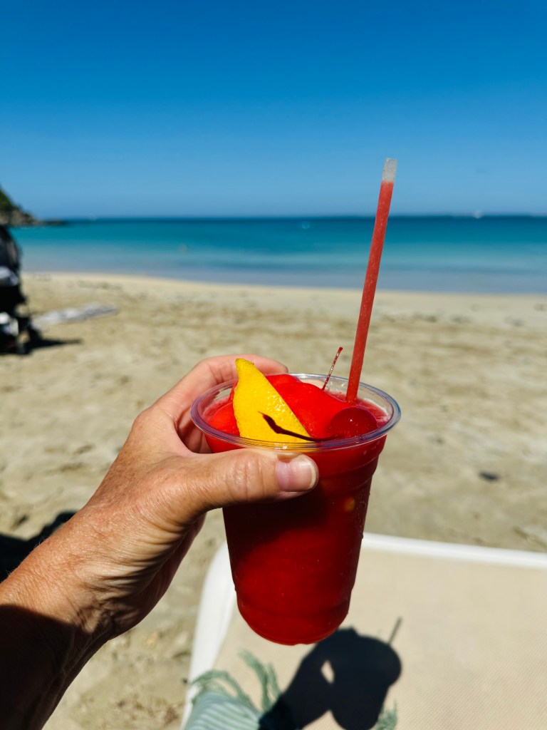 En hand som håller en drink med röd färg och fruktskivor på stranden, med havet i bakgrunden.