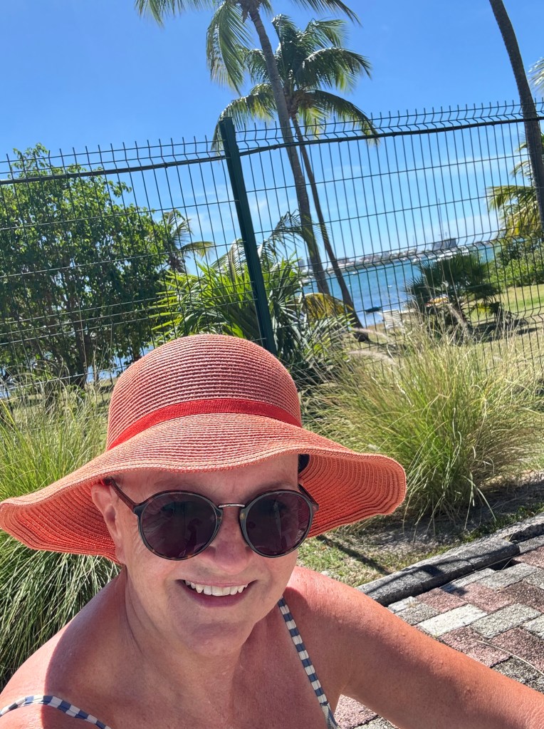 En leende kvinna i solhatt och solglasögon sitter vid en pool med palmträd och havet i bakgrunden.