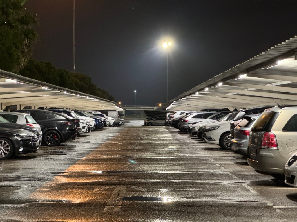 En parkeringsplats med bilar under tak i nattbelysning, reflektioner av regn på marken.