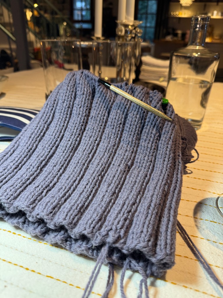 En grå ribbad stickning med en virknål, placerad på ett bord med flera föremål i bakgrunden.